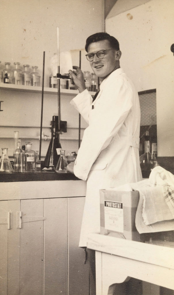 vintage photograph of a man in a lab suit holding a beaker in front of chemistry lab.